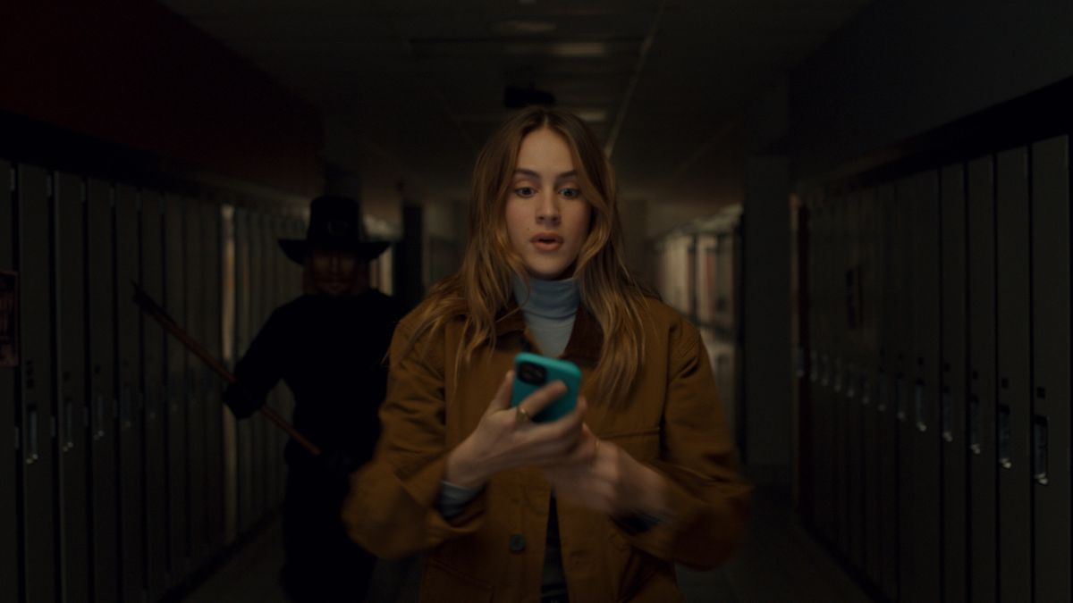 En kvinna (Nell Verlaque) går i en korridor på sin telefon medan en man i mask som håller en yxa följer efter henne.