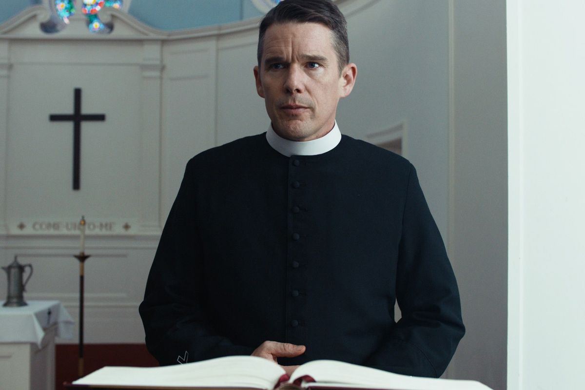 En man i prästdräkt (Ethan Hawke) står bakom en talarstol med ett svart kors synligt i bakgrunden.
