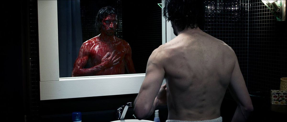 En man står framför sin badrumsspegel och kollar hans bröst, som är täckt av blod i Chimeres