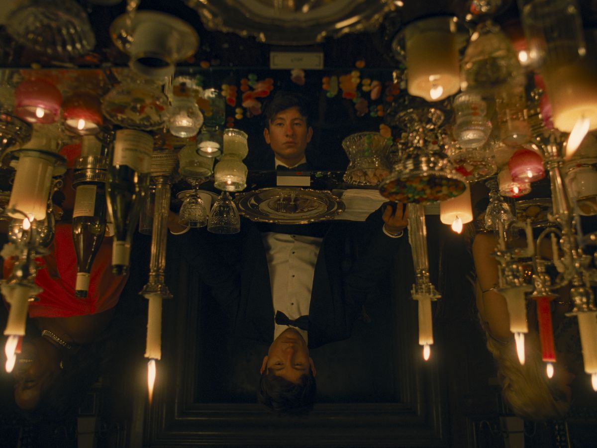 Oliver (Barry Keoghan), in black tie dress, sits at what appears to be an fancy table covered in candles of all descriptions, reflecting his face back at him — except the more you look, the more it’s clear that the reflection is in a different position, standing with its eyes lowered. From the movie Saltburn