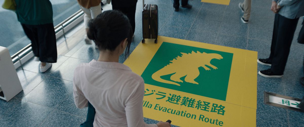 En kvinna står på en flygplats framför en skylt på golvet som markerar en evakueringsväg från Godzilla i Apple TV Plus-programmet Monarch: Legacy of Monsters