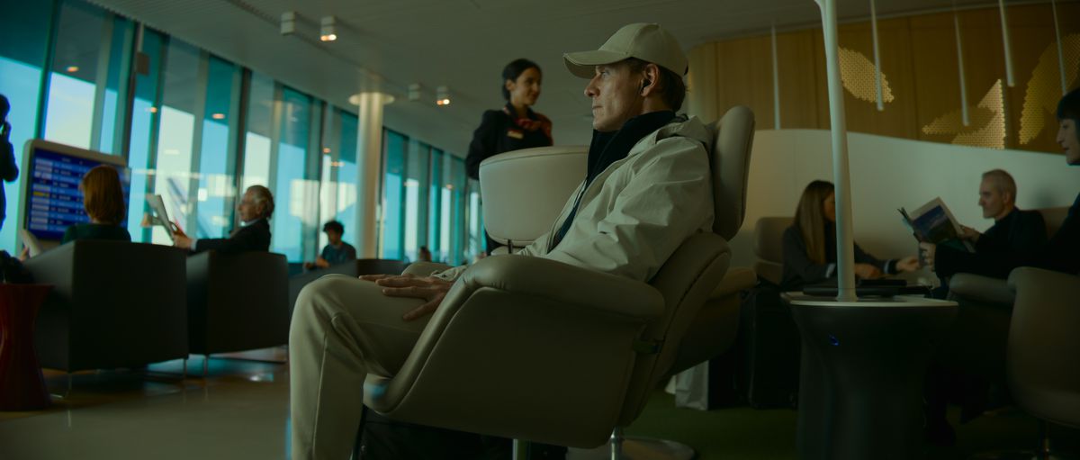 Michael Fassbender som Mördaren, sitter i ett väntrum på flygplatsen, bär en basebollkeps, ser uttråkad ut