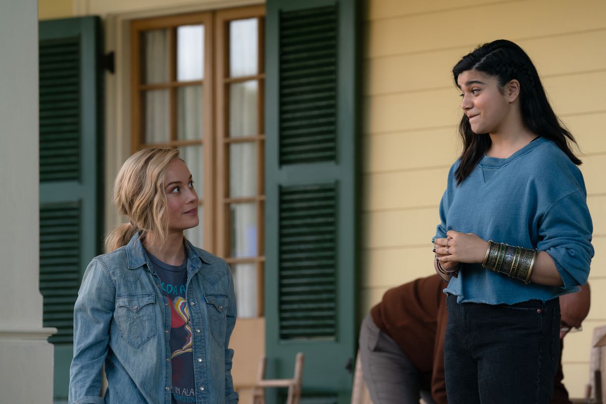 Brie Larson som Captain Marvel/Carol Danvers och Iman Vellani som Ms. Marvel/Kamala Khan står utomhus på en veranda och ler mot varandra i Marvel Cinematic Universe-filmen The Marvels
