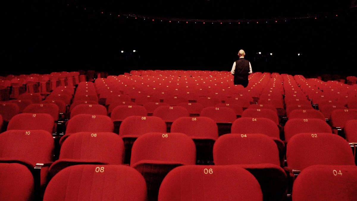 Robert Fripp, wearing a vest, has his back to the camera and is surrounded by empty red audience chairs in In the Court of the Crimson King: King Crimson at 50.