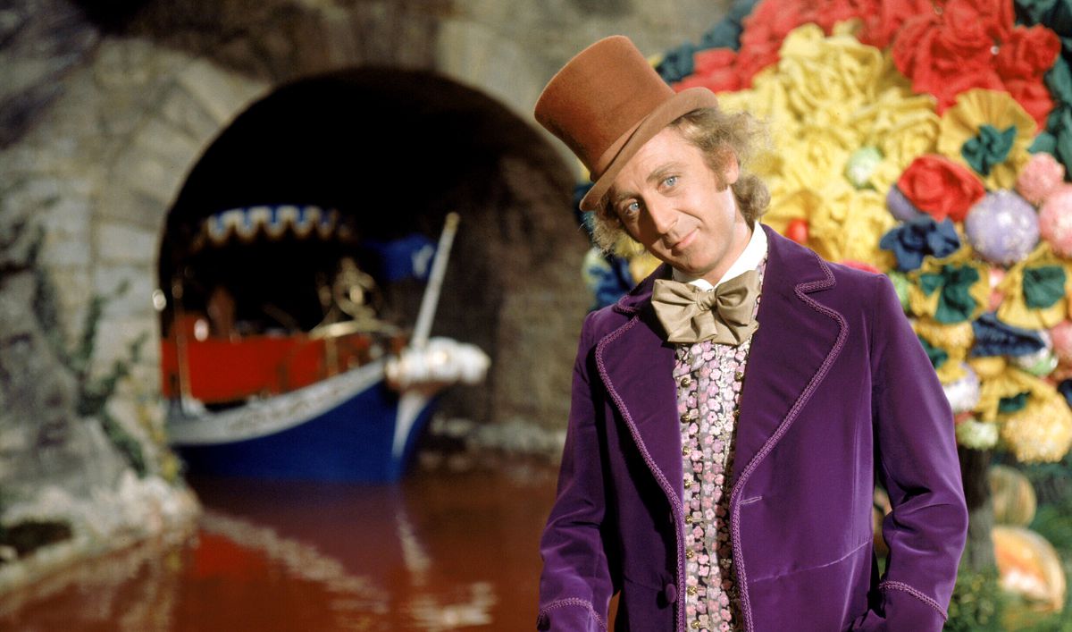 Willy Wonka (Gene Wilder, in purple velvet jacket and tan top hat) stands in front of a chocolate river as a ship appears out of a tunnel behind him in 1971’s Willy Wonka & the Chocolate Factory