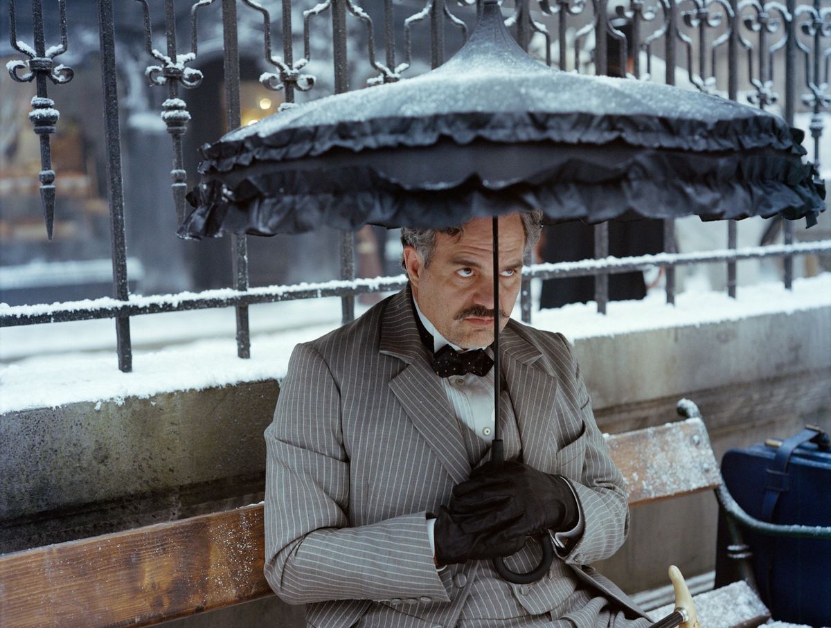 Duncan Wedderburn (Mark Ruffalo, in pinstriped grey suit) pouts theatrically under a frilly grey parasol while sitting on a wooden bench in the snow in Poor Things