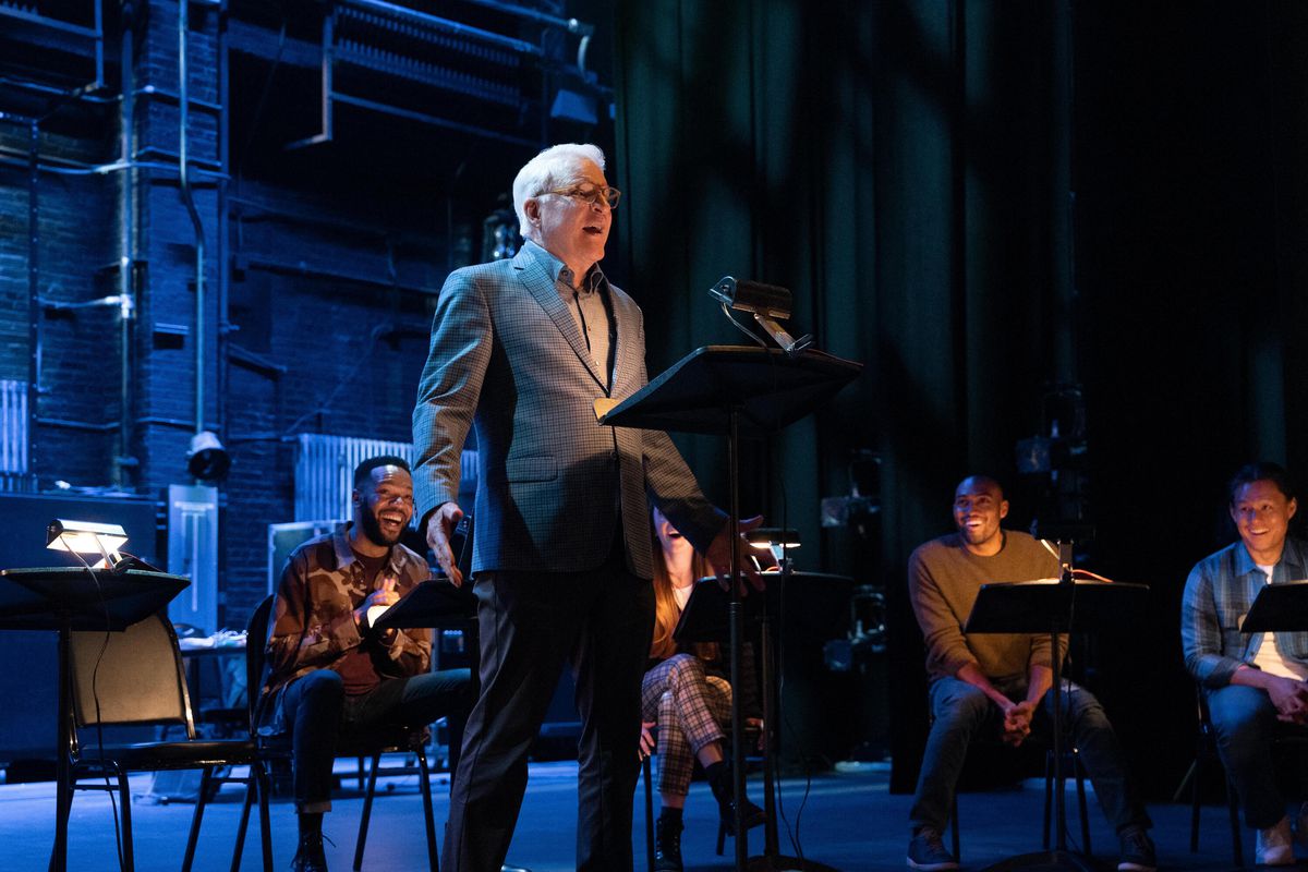 Steve Martin standing and singing in front of a music stand with people seated behind him on a theater stage in Only Murders in the Building