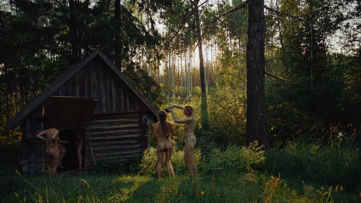 Fyra nakna kvinnor står runt eller inne i en liten bastu gjord av råa stockar, i en livligt grön skog, i Smoke Sauna Sisterhood