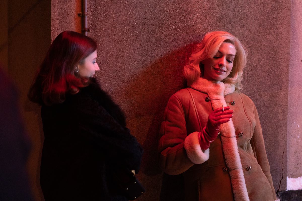 Anne Hathaway, i blond peruk och shearling rock, röker lutad mot en neon-dränkt vägg som Rebecca medan Thomasin McKenzie tittar på i filmen Eileen.