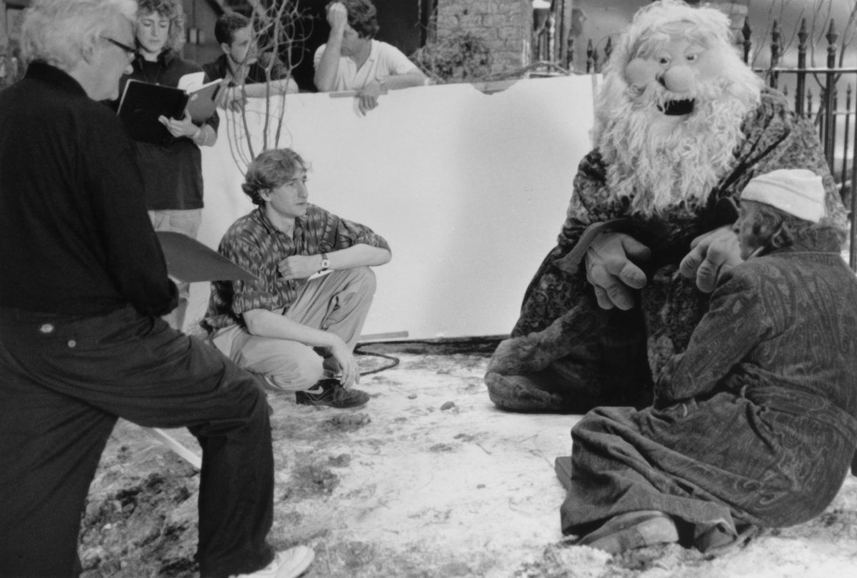 LtR: Various crew, Brian Henson (director), the Ghost of Christmas Present, and Michael Caine costumed as Ebeneezer Scrooge crouch, sit, and stand on a set strewn with fake snow, a giant white reflective board and other filmmaking gear visible.