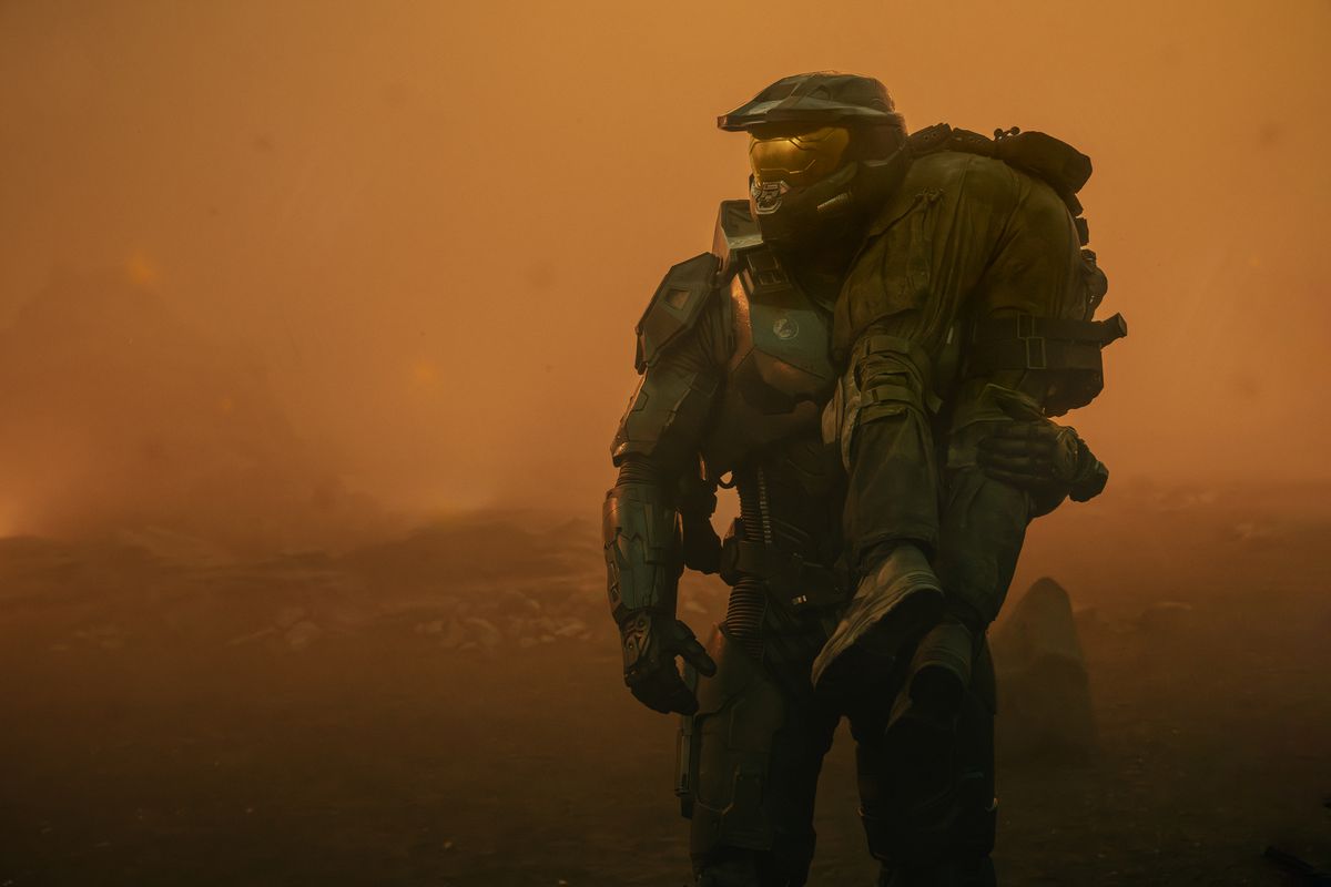 Master Chief holds as lumped body over his shoulder while walking through an orange-tinged desert landscape in Halo season 2