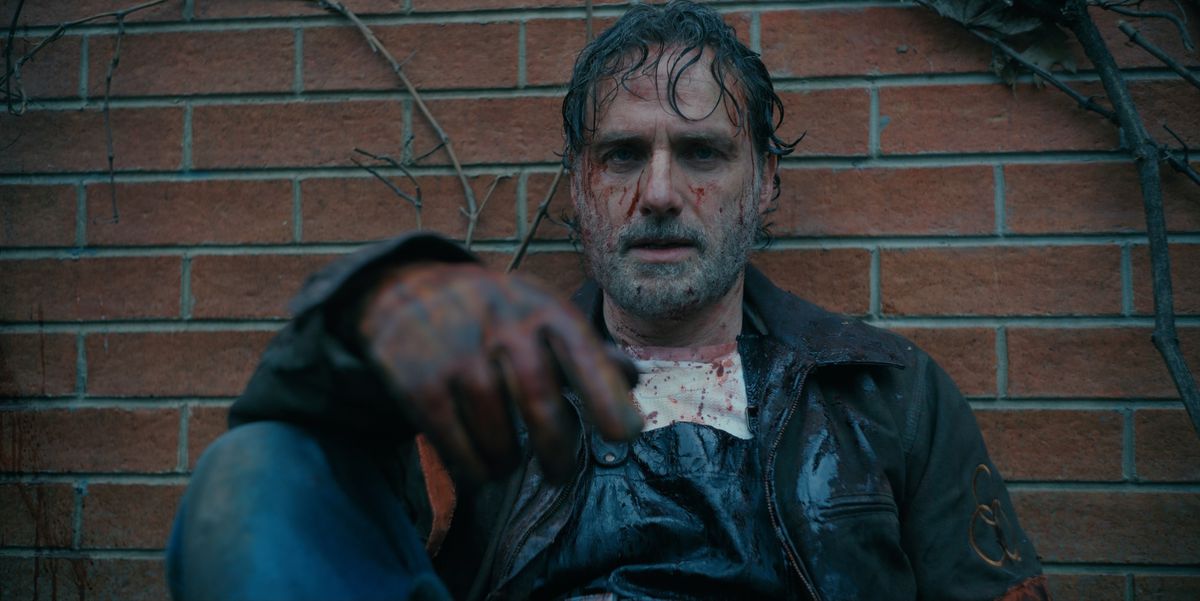 Andrew Lincoln as Rick Grimes with a bloody face, sitting against a brick wall in The Walking Dead: The Ones Who Live