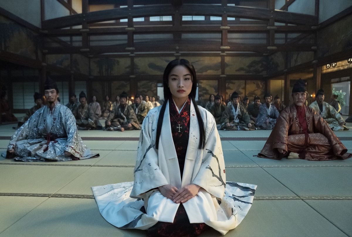 Anna Sawai, wearing white, kneels in the Shōgun TV show. Many other people sit cross-legged behind her.