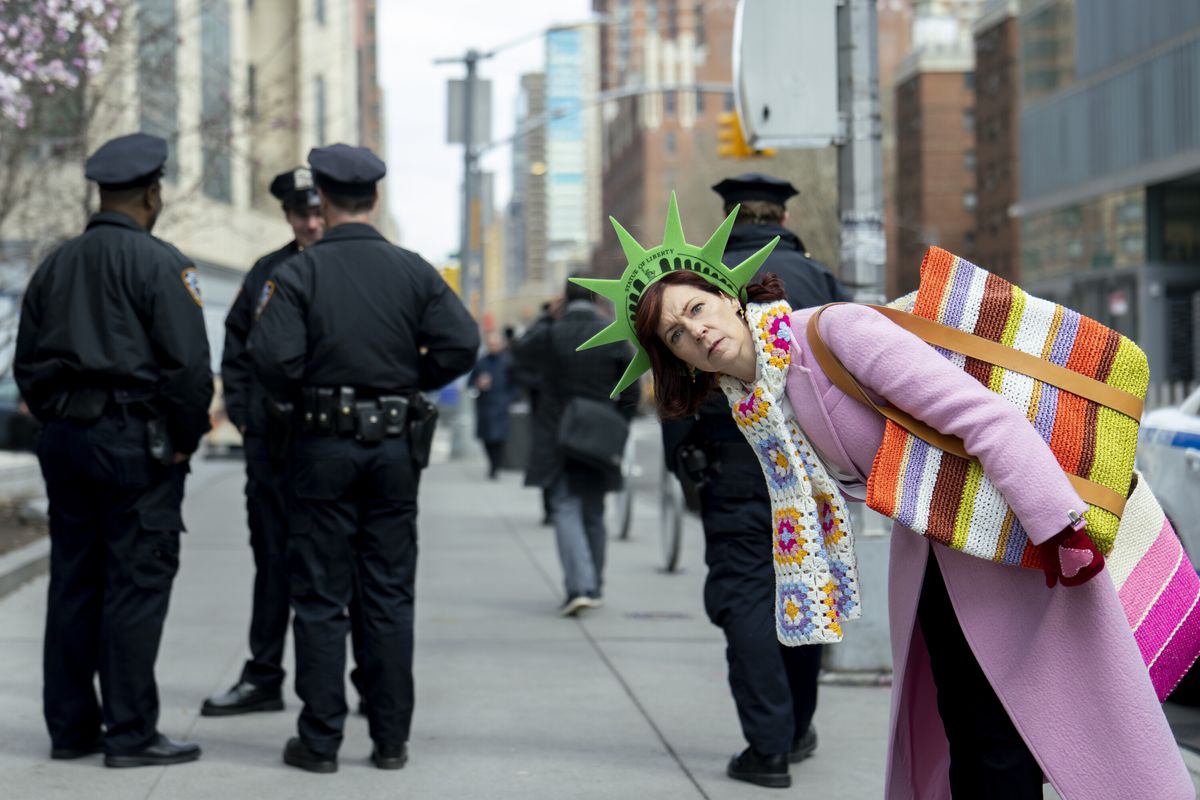 Carrie Preston as Elsbeth Tascioni, wearing a green felt statue of liberty hat, a pink/purple coat and a flashy knitted scarf + bag combo, leans over and looks towards the camera quizzically in Elsbeth