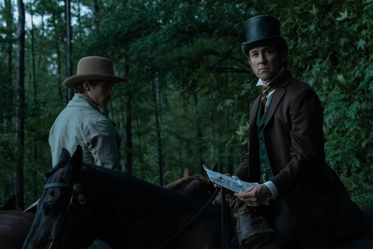 Tobias Menzies and Brandon Flynn ride horses and wear hats in the forest in Manhunt. Menzies holds what looks like a letter or a map