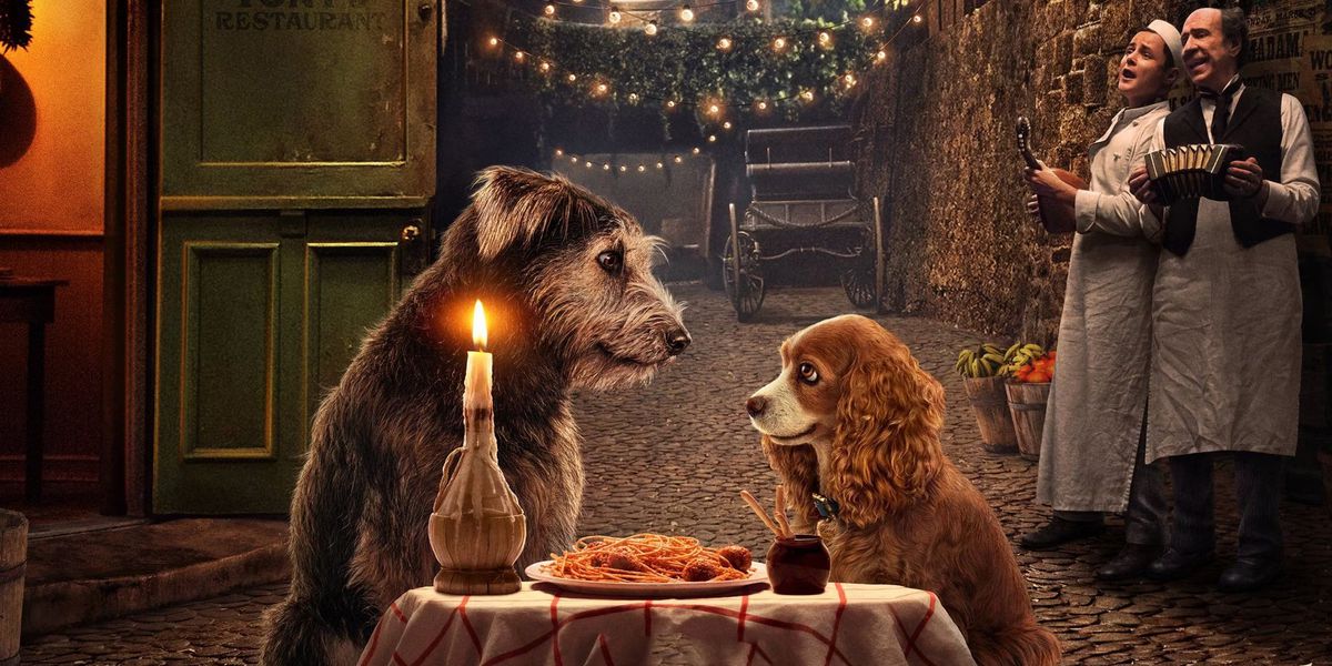 Tramp (a Schnauzer-mix dog) and Lady (a Cocker Spaniel) sit across from each other at a table sporting a candle in a wine bottle and a plate of spaghetti and meatballs, while two men in chefs’ whites, one with a lute and one with an accordion, serenade them in a promo image from Disney’s live-action Lady and the Tramp