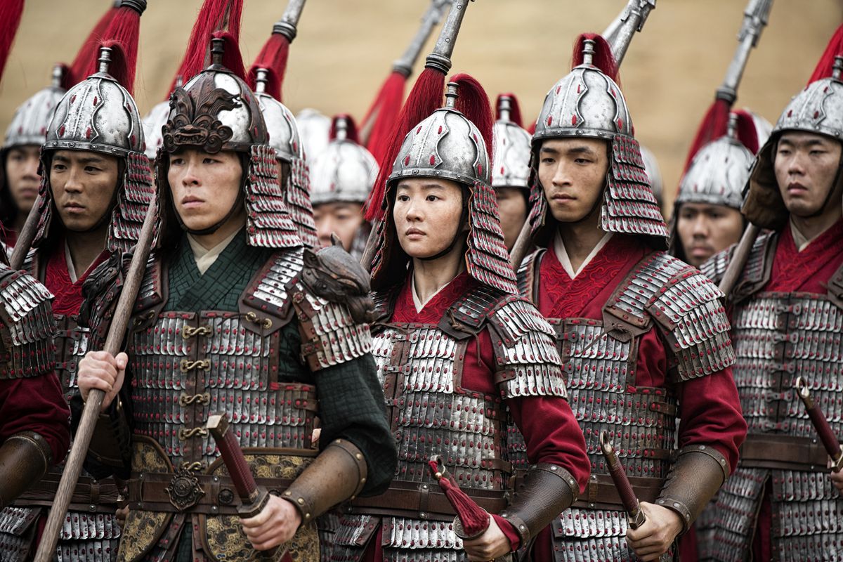 Mulan (Liu Yifei), disguised as a boy in metal lamellar armor and metal helmet, stands among a group of identically dressed soldiers in the 2020 live-action Mulan remake