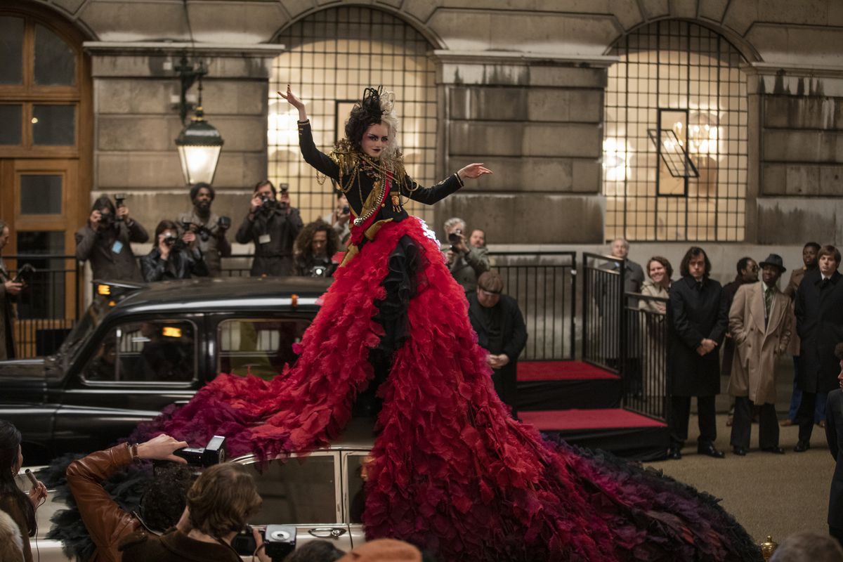 Cruella de Vil (Emma Stone) in a psuedo-military-style blouse decked with medals and epaulettes and a tumbling, fluffy red skirt stands on top of a car, posing for photographers and a crowd, in the 2021 live-action movie Cruella