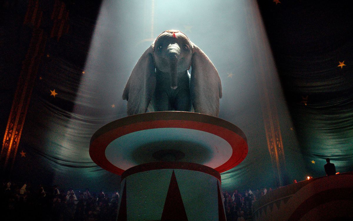 CG Dumbo the elephant stands on a high red-and-white platform in a bright spotlight at the circus as a dimly seen audience watches in the 2019 live-action Dumbo remake