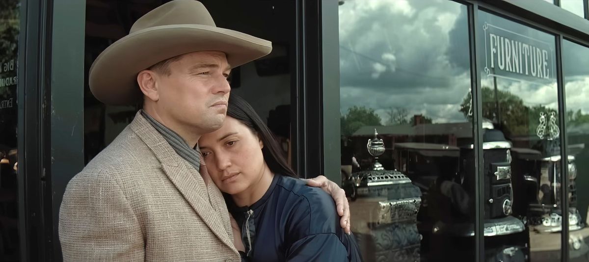 Ernest (Leonardo DiCaprio, i brun kostym och cowboyhatt) tröstar sin fru Mollie (Lily Gladstone) när hon lutar sig mot honom framför ett möbelfönster i Killers of the Flower Moon