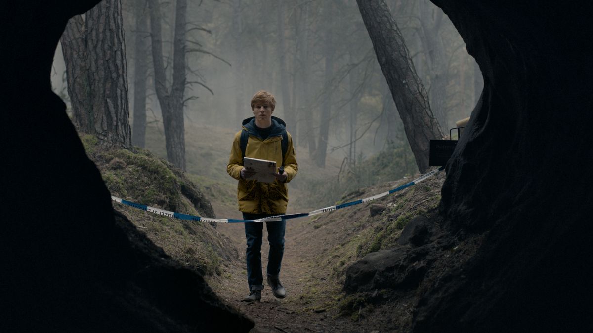 En ung man klädd i en gul regnrock står framför den avdelade ingången till en stor grotta i en skog i mörkret.