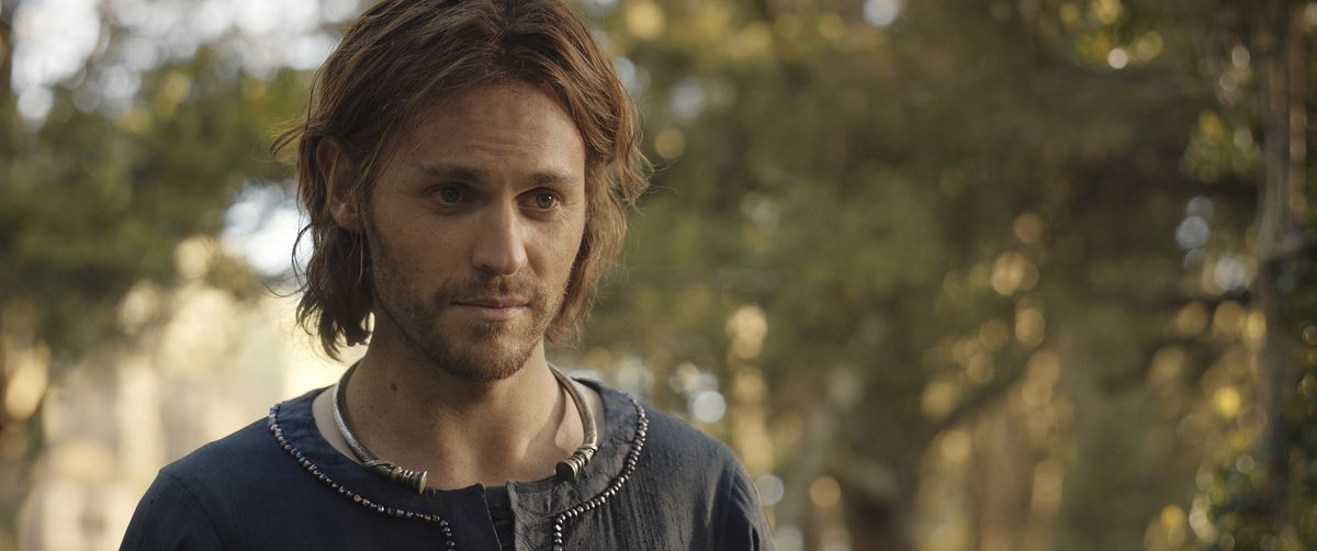 Charlie Vickers as Halbrand, wearing a blue tunic and making your standard solemn heartthrob face while standing amongst some trees in Lord of the Rings: The Rings of Power