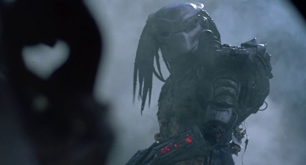En bepansrad utomjordisk krigare som bär en mask med dreadlocks står i en djungel omgiven av rök i Predator.