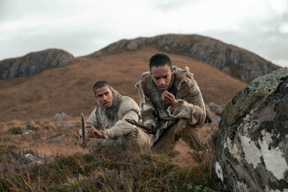 Två paleolitiska män, Adem och Geirr (Chuku Modu och Kit Young) håller i sina spjut när de spårar något över en brun, gräsbevuxen, stenig kulle i Out of Darkness