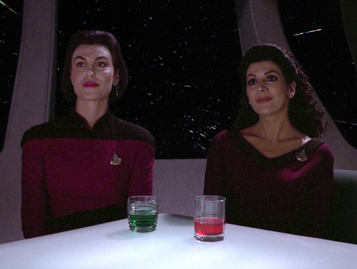 Ensign Ro Laren and Counselor Troi sit next to each other at a bar table, smiling awkwardly and perhaps angrily in Star Trek: The Next Generation. 