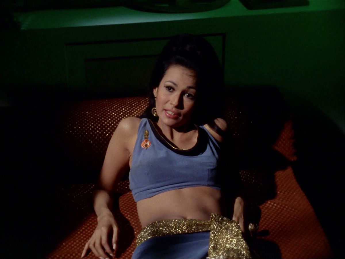 A woman loungeds on a bed in a Star Fleet-like uniform, but it has been altered to be a crop top, and given a glittery gold sash at the waist. 