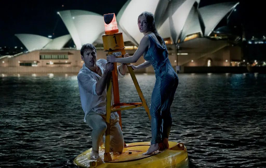 En man och en kvinna klamrar sig fast vid en boj i en vik med Sydney Opera House synligt i bakgrunden.