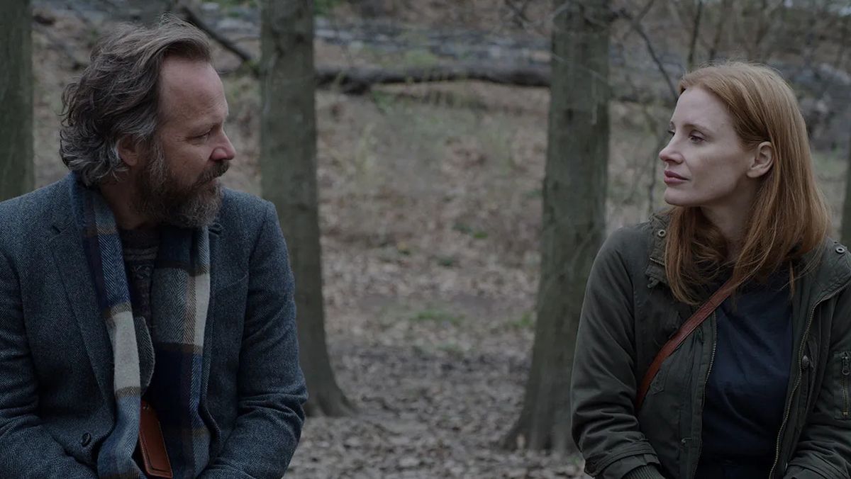 A bearded man and a red-haired woman sit beside one another in a forest.