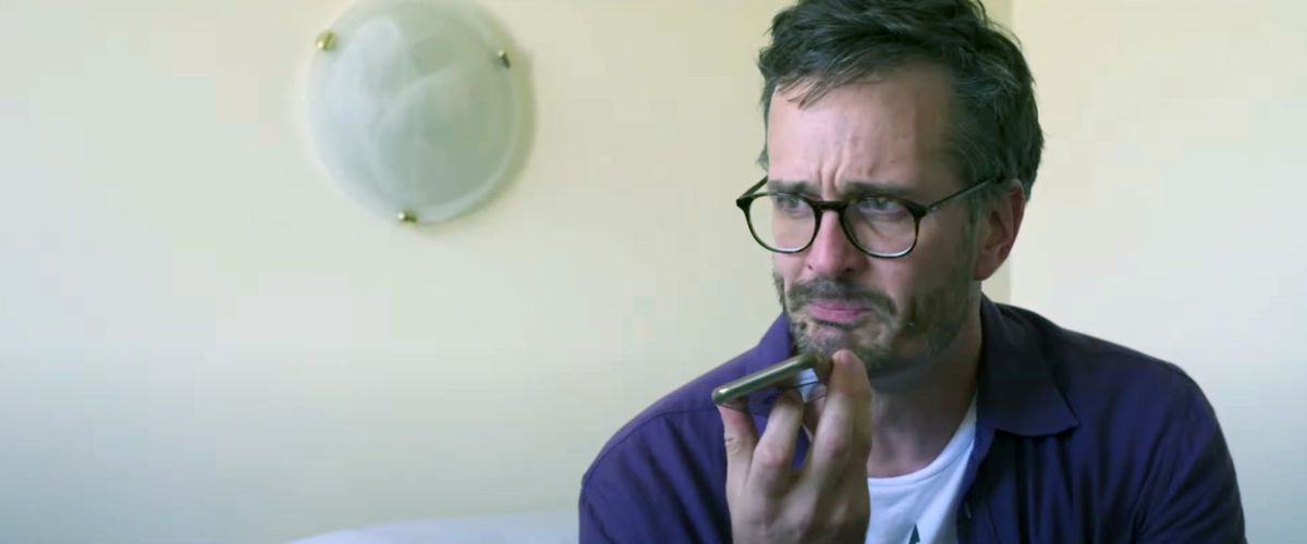 Journalisten David Farrier sitter på en säng i ett rum med vita väggar och håller en mobiltelefon under hakan och gråter i en scen från sin dokumentär Mister Organ