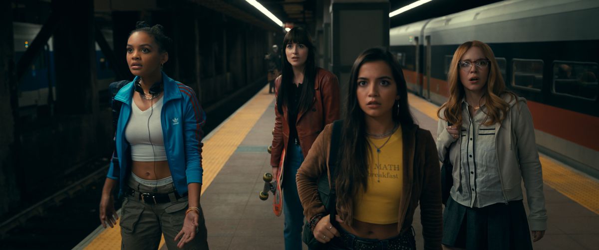 Tre unga kvinnor står på en tunnelbaneplattform och ser chockade ut.  En vuxen kvinna står lite bakom dem, på vakt. 