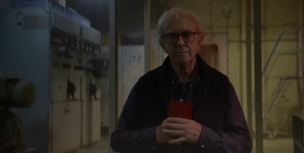 Jonathan Pryce standing toward the screen with glasses on and a sweater while clutching a book in 3 Body Problem