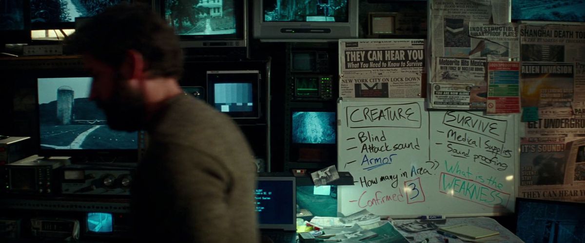John Krasinski sitter oskarp vid en dator, kameran visar oss en whiteboard med massor av ledtrådar till utomjordingarna och tidningsklipp om invasionen, i A Quiet Place