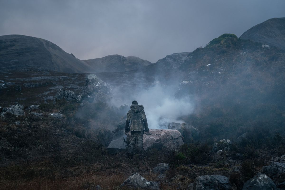 En person klädd i grova, handgjorda byxor och kavaj i grå päls och läder står med ryggen mot kameran, vänd mot en dimmig dal mellan två steniga kullar i Out of Darkness