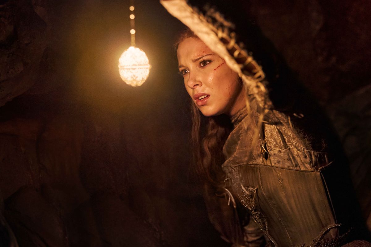 Millie Bobby Brown in Netflix’s Damsel stands in a cave with a bow on her back