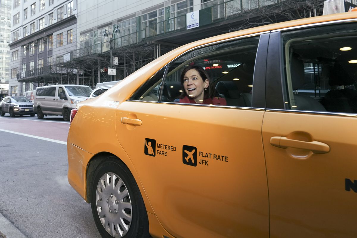 Elsbeth sitter bak i en taxi och ler ut genom fönstret i en stillbild från Elsbeth