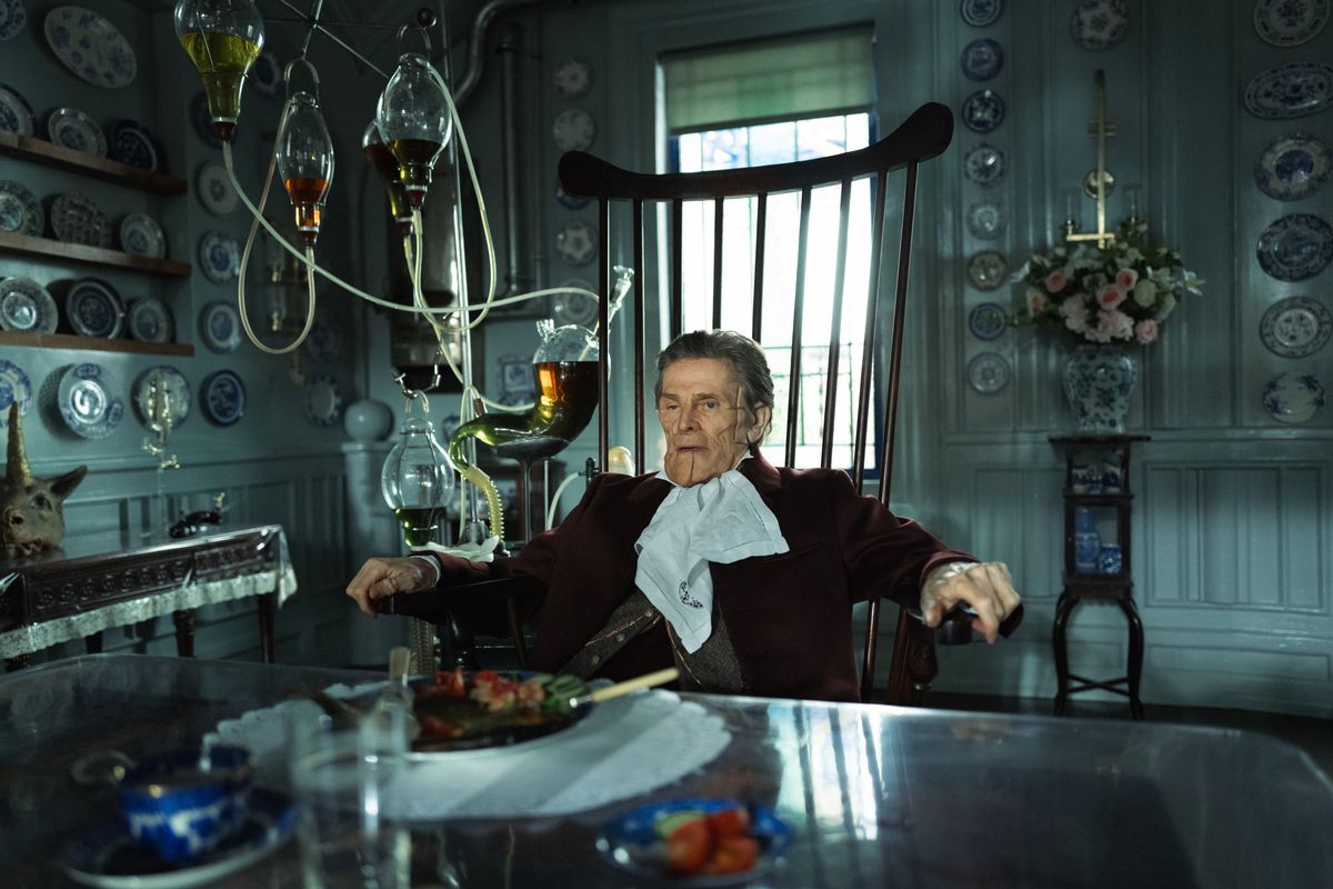 Willem Dafoe, med ett ärrat ansikte, sitter på en högryggad stol med en glasapparat som innehåller olika vätskor bredvid sig