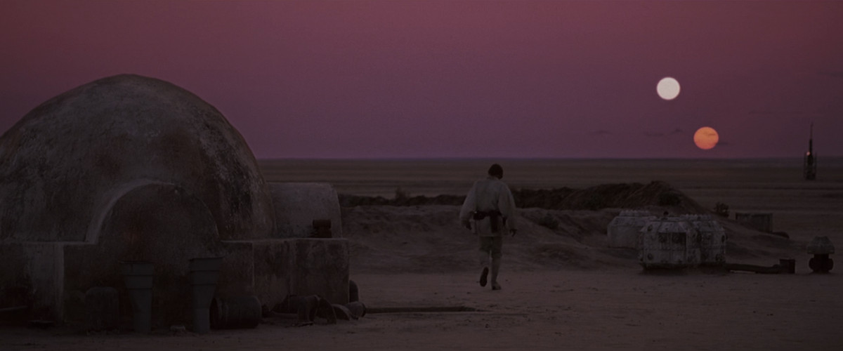 Luke Skywalker går över sin sandiga gård mot horisonten, två solar går ner på himlen. 