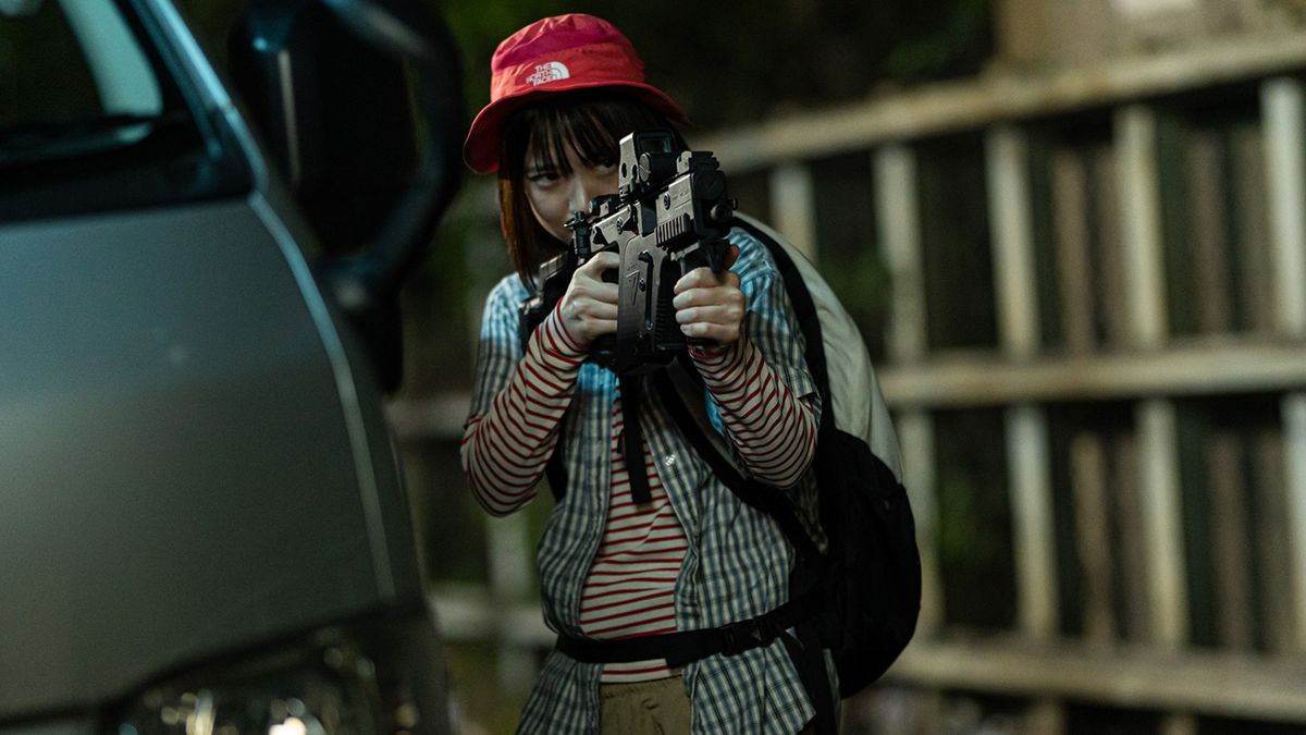 En ung flicka som bär en röd bucket-hatt och en ryggsäck håller och siktar en maskingevär i Baby Assassins 2 Babies.