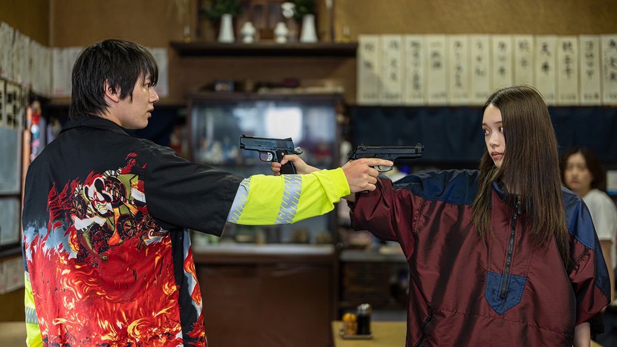Akari Takaishi och en ung man i en grymt högljudd jacka riktar pistoler mot varandra inomhus i Baby Assassins 2 Babies.
