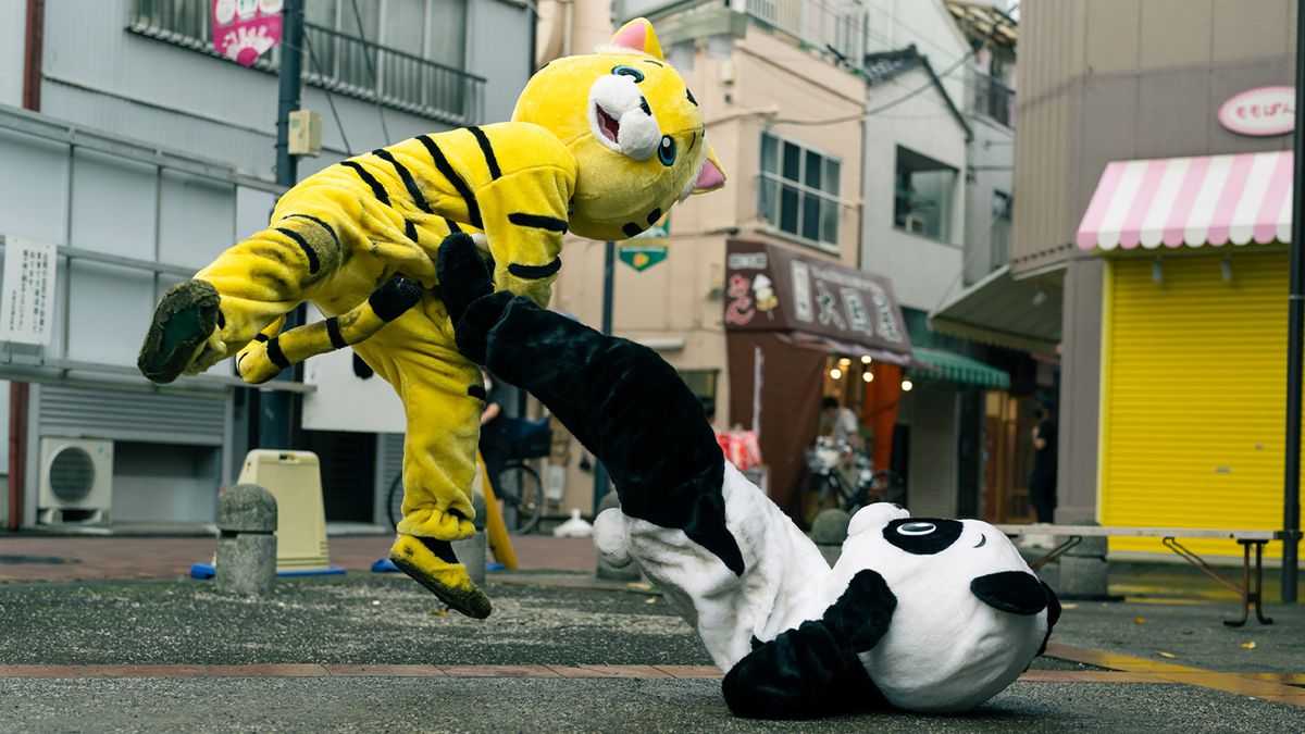 Ett slagsmål mellan två människor i stora djurkostymer, när en panda som sitter på marken sparkar en hoppande tiger i magen, i Baby Assassins 2