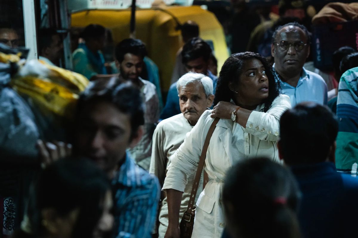 A woman wearing a white jacket and purse gazing off-screen while standing in a crowd of people.