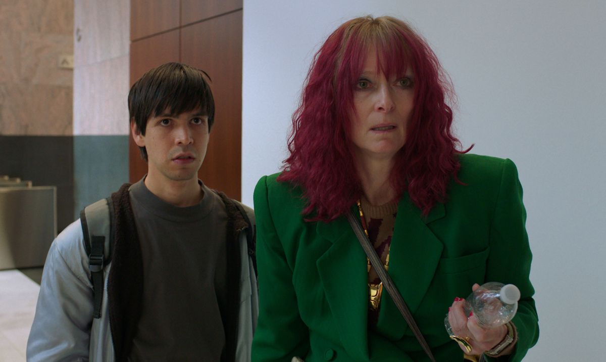 Tilda Swinton in a green blazer and fuschia hair holds a water bottle, addressing someone offscreen with Julio Torres in a hoodie and backpack standing behind her, from Problemista