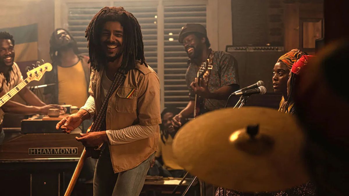A smiling man with dreadlocks standing next to a band of musicians playing.
