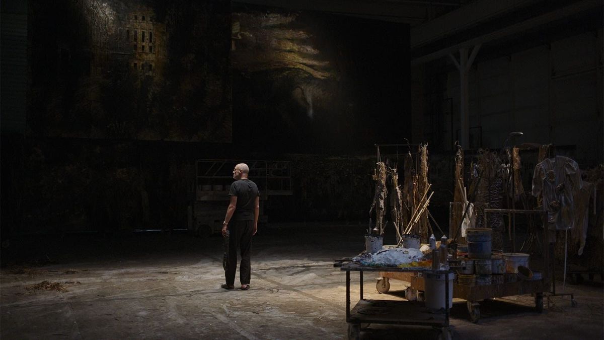 En man som står i en mörk ateljé omgiven av konstförnödenheter och stora målningar.