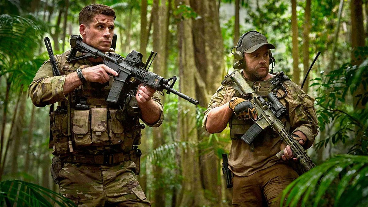 Två män i armékamouflage som håller gevär i en tropisk skog.