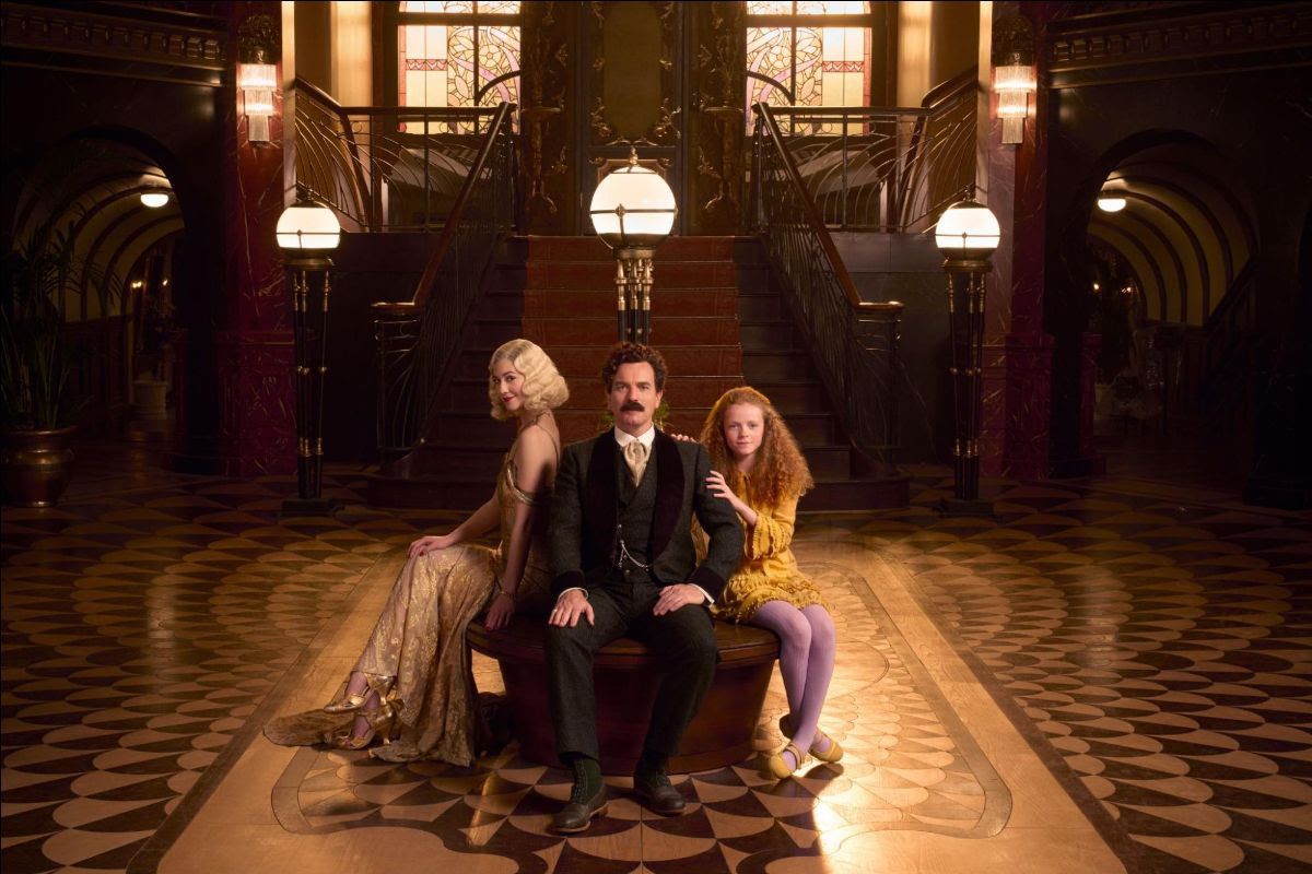 Ewan McGregor, Mary Elizabeth Winstead, and Alexa Goodall sit on a round table, on top of a fancy floor, in front of an ornate staircase in A Gentleman in Moscow. 